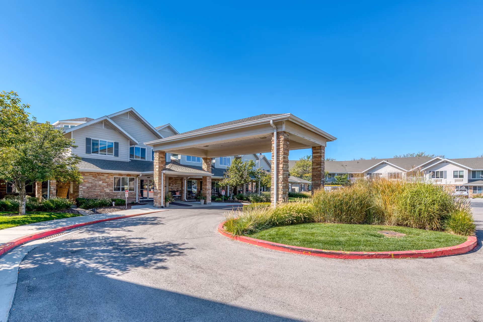 Exterior view of Solstice Senior Living at Sandy facility showing the entrance with a covered drop-off area supported by stone pillars, surrounded by landscaped greenery and a clear blue sky.