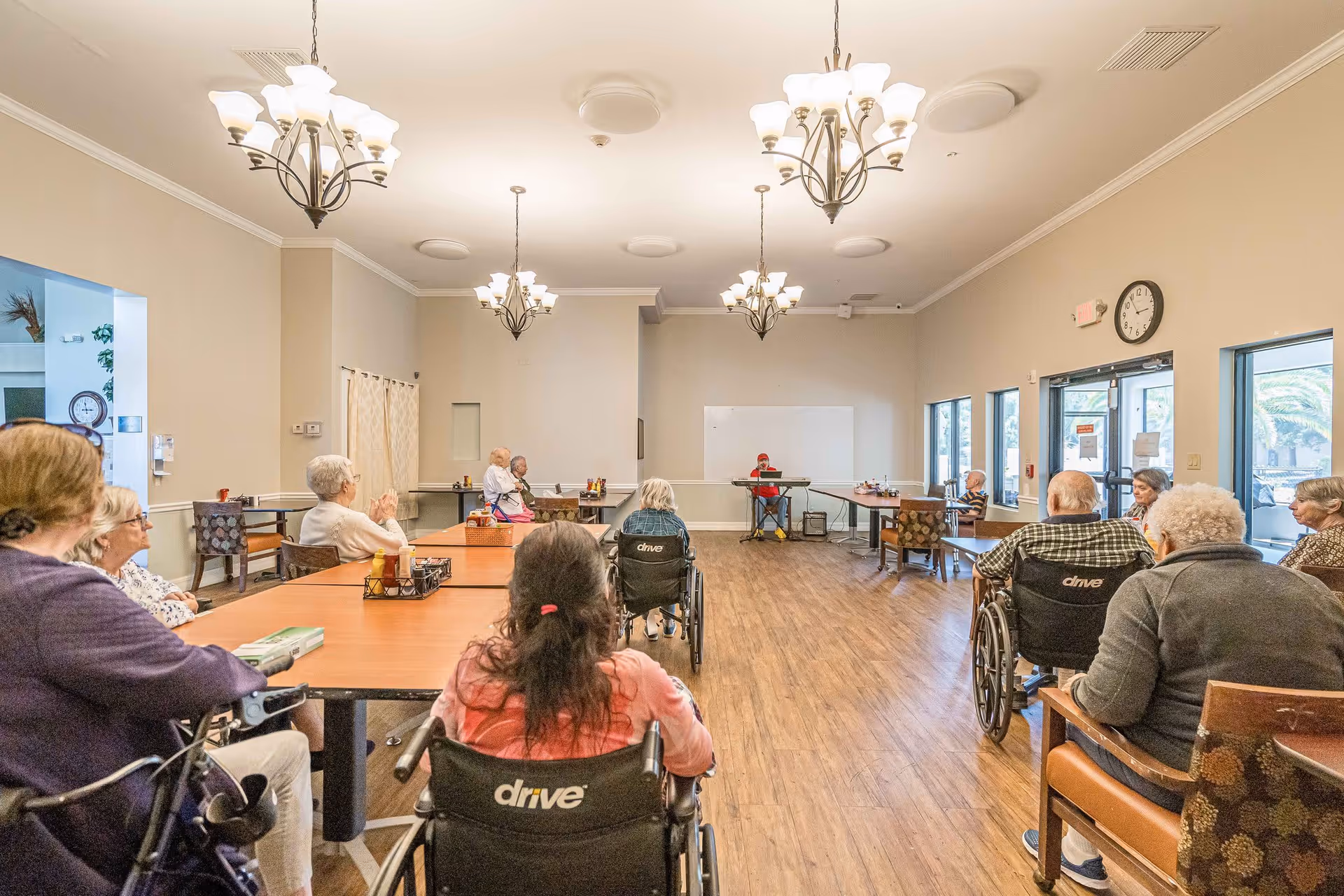 A group of elderly people, some in wheelchairs, seated around tables in a large, well-lit room with wooden floors and chandeliers. A person at the front appears to be leading an activity or presentation. The room has large windows and a clock on the wall.