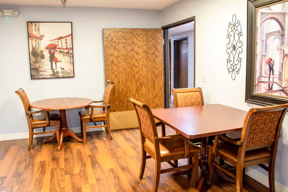 Interior room with two wooden tables and six chairs on a wood floor. One table is round with two chairs, and the other is square with four chairs. The walls are light-colored with two framed paintings and a decorative metal wall piece. A wooden door is partially open, revealing a hallway.