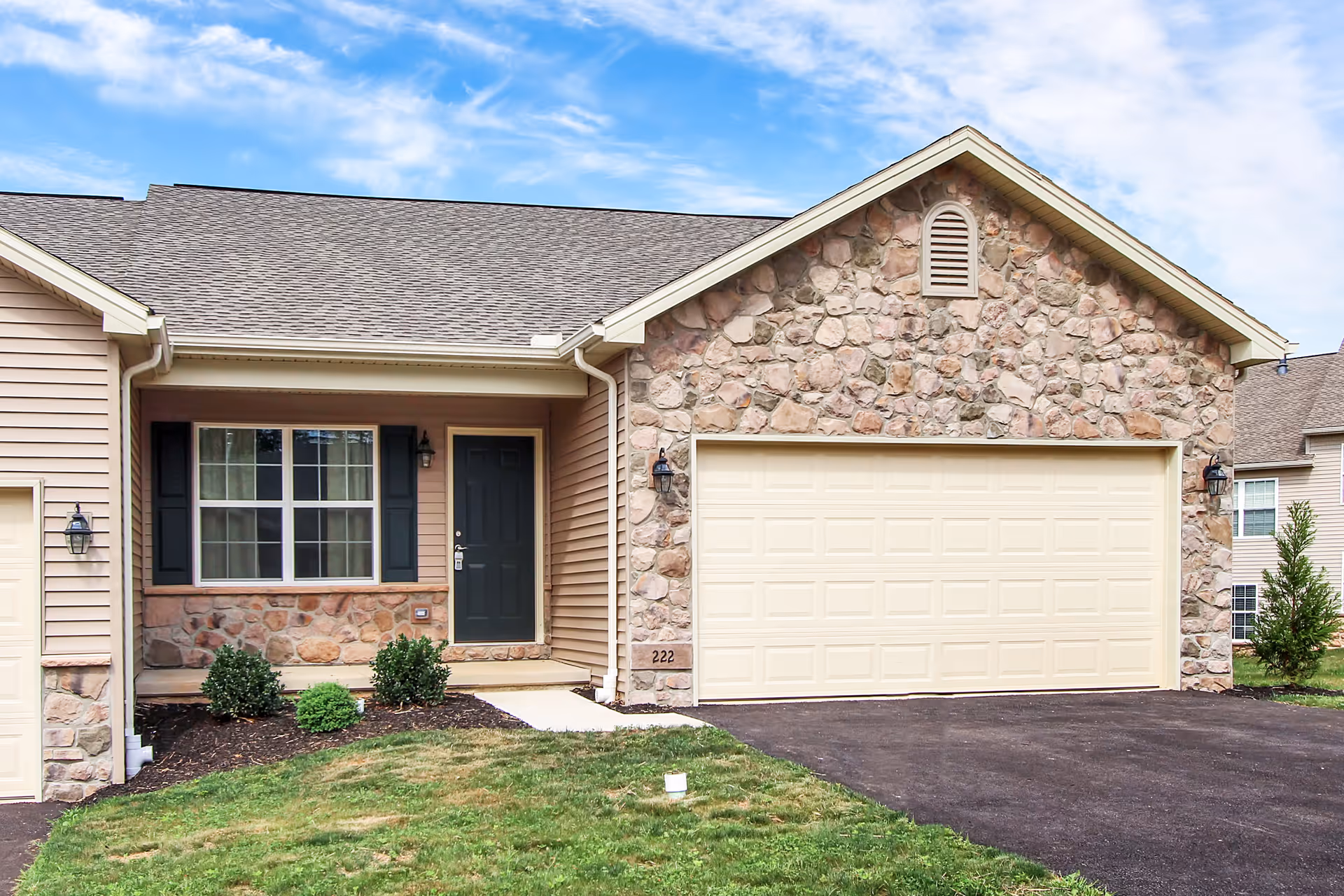 Front exterior view of a single-story residential building with a stone and siding facade, a black front door, a large window with black shutters, a two-car garage with a cream-colored door, and a small landscaped area with bushes and grass.