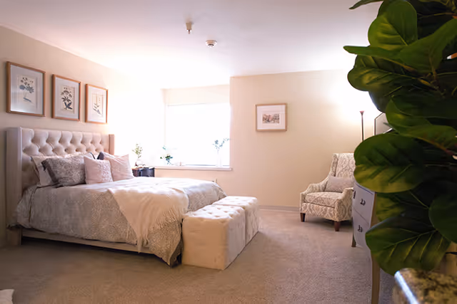 A cozy bedroom in a senior living facility featuring a large bed with a tufted headboard, multiple pillows, and a light blanket. There is a cushioned bench at the foot of the bed, a patterned armchair in the corner, a floor lamp, framed artwork on the walls, a window letting in natural light, and a large green plant partially visible in the foreground.