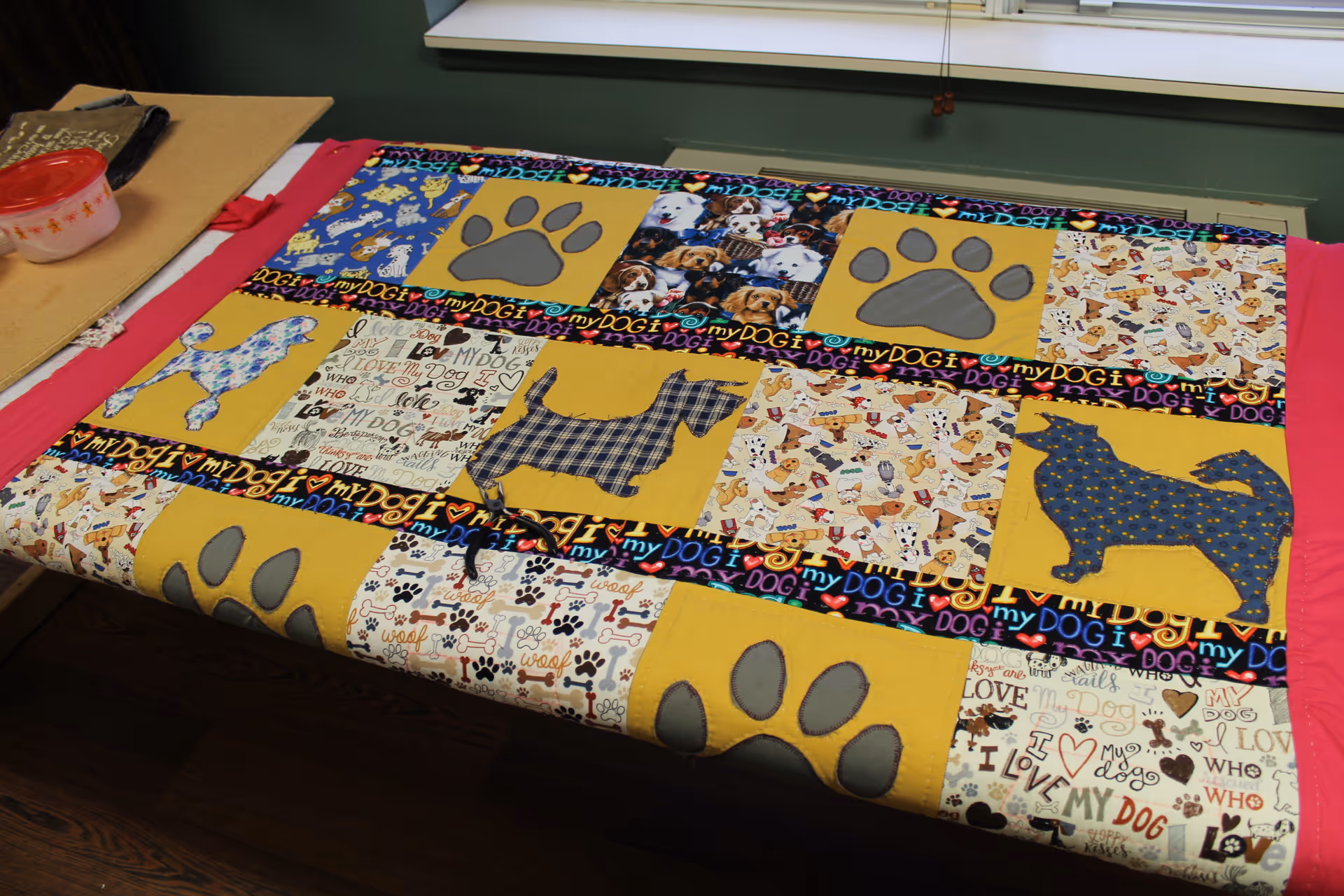 A colorful quilt with various dog-themed patterns and designs, including paw prints and different dog silhouettes, laid out on a table with a pink border around the quilt.