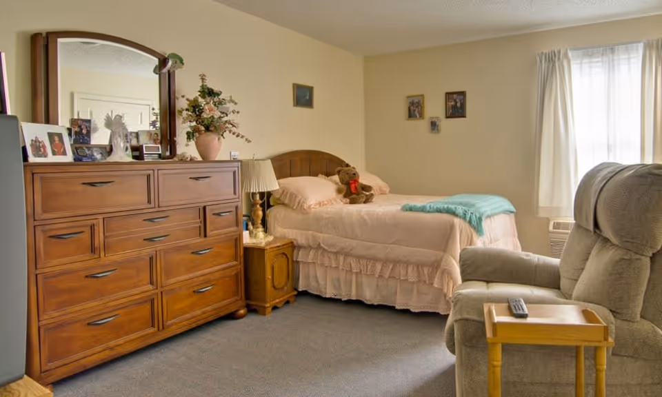 A cozy senior living bedroom with a wooden dresser topped with framed photos and a vase of flowers, a bed with pink bedding and a teddy bear, a small wooden nightstand with a lamp, a comfortable recliner chair with a small wooden side table holding a remote control, and a window with white curtains letting in natural light.