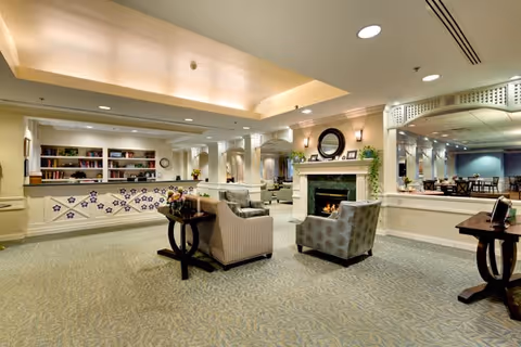 A spacious, well-lit common area with comfortable seating including armchairs and sofas arranged around a fireplace. The room features a reception desk with shelves behind it, decorative plants, a large mirror above the fireplace, and a carpeted floor with a subtle pattern. The ceiling has recessed lighting and a tray design with soft lighting.