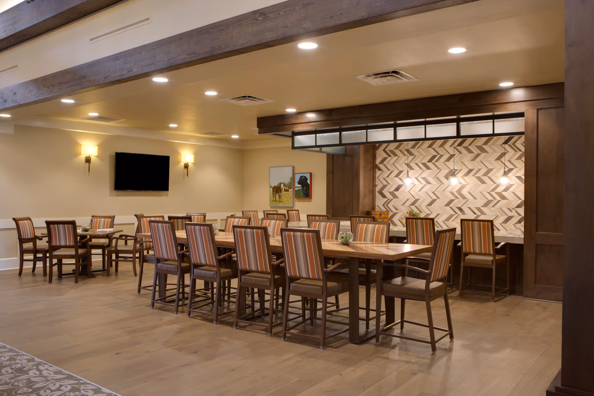 A spacious dining room with multiple wooden tables and chairs featuring striped upholstery. The room has warm lighting with wall sconces and recessed ceiling lights. A flat-screen TV is mounted on the wall, and there are two framed pictures of dogs. The back wall has a decorative chevron pattern with three pendant lights hanging above a counter area.