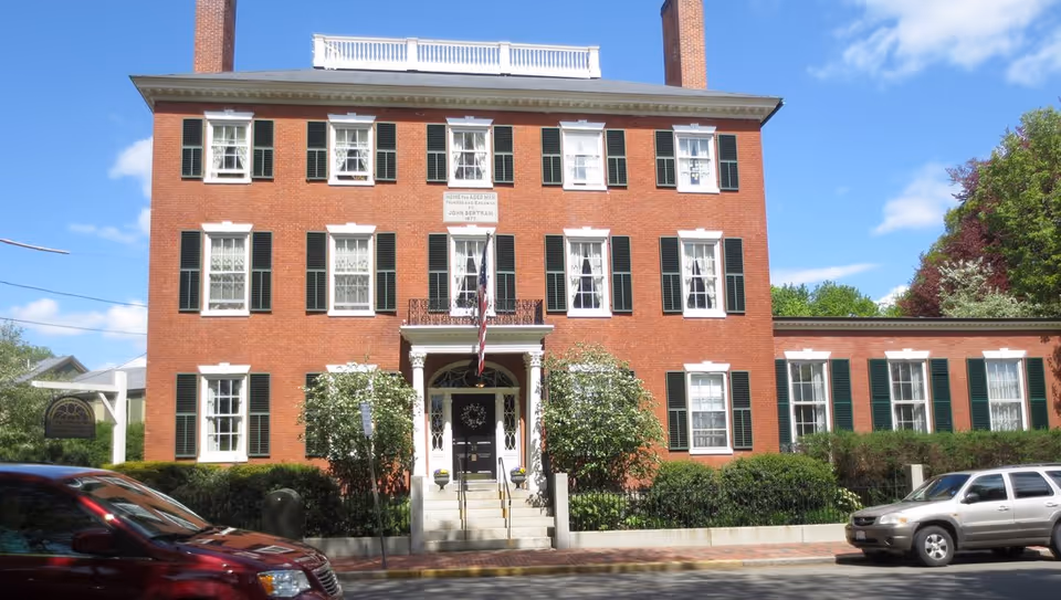 Front exterior of a three-story red brick building with green shutters, an American flag at the entrance, and cars parked on the street.