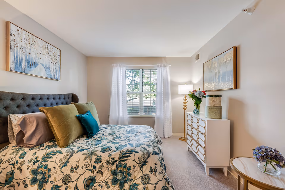 A cozy bedroom with a bed featuring a tufted dark gray headboard, floral patterned bedding in shades of blue and green, and multiple pillows in green and teal. There is a window with white curtains letting in natural light. On the right side, there is a white dresser with a geometric pattern, a vase with flowers, and decorative boxes. A floor lamp with a gold stand is next to the dresser. Two framed artworks hang on the walls, one above the bed and one above the dresser. A small round side table with a glass vase holding purple flowers is also visible.