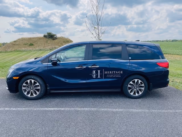 A dark blue minivan parked on a paved road with grassy fields and a partly cloudy sky in the background. The minivan has the Heritage Hills Retirement Home logo and text on the side.