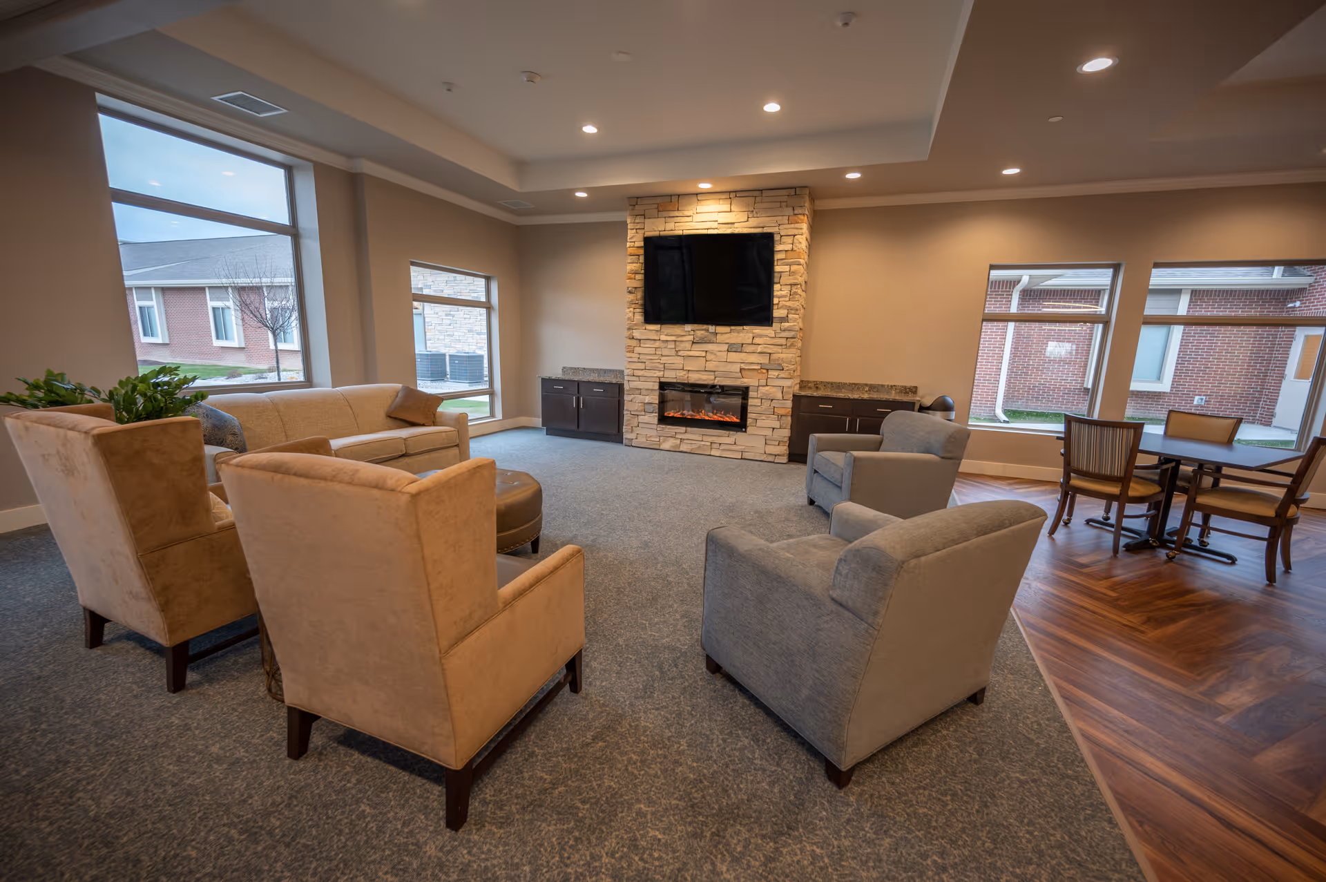A cozy living room area with beige and gray armchairs arranged around a stone fireplace with a mounted flat-screen TV above it. Large windows let in natural light, and a small dining table with chairs is visible in the background on a wooden floor section.
