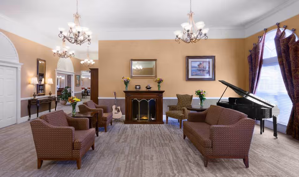 A cozy living room area with brown upholstered chairs and a sofa arranged around a wooden fireplace. A guitar leans against the wall next to the fireplace. There is a grand piano near a large window with purple curtains. The walls are painted beige with white trim, and the room is illuminated by elegant chandeliers and wall lamps. A console table with lamps and a mirror is visible on the left side.