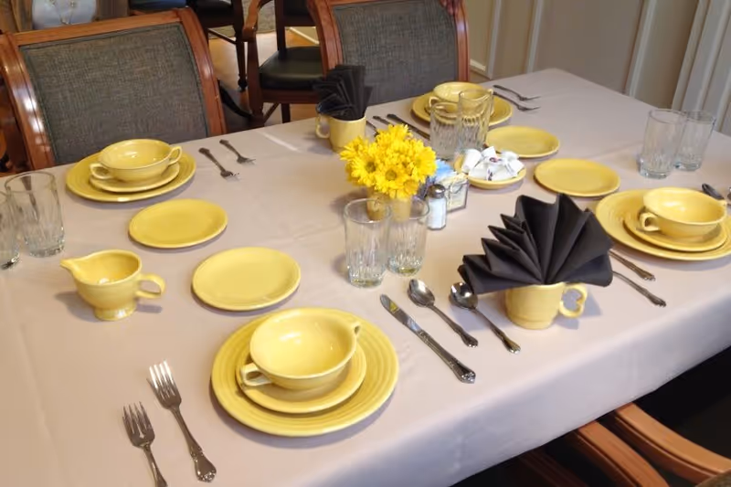 A dining table set for six with yellow dishes, cups, and saucers on a white tablecloth. Each place setting includes a fork, knife, spoon, and glass. Two yellow mugs hold black folded napkins, and a small vase with yellow flowers is in the center of the table along with salt and pepper shakers and sugar packets.