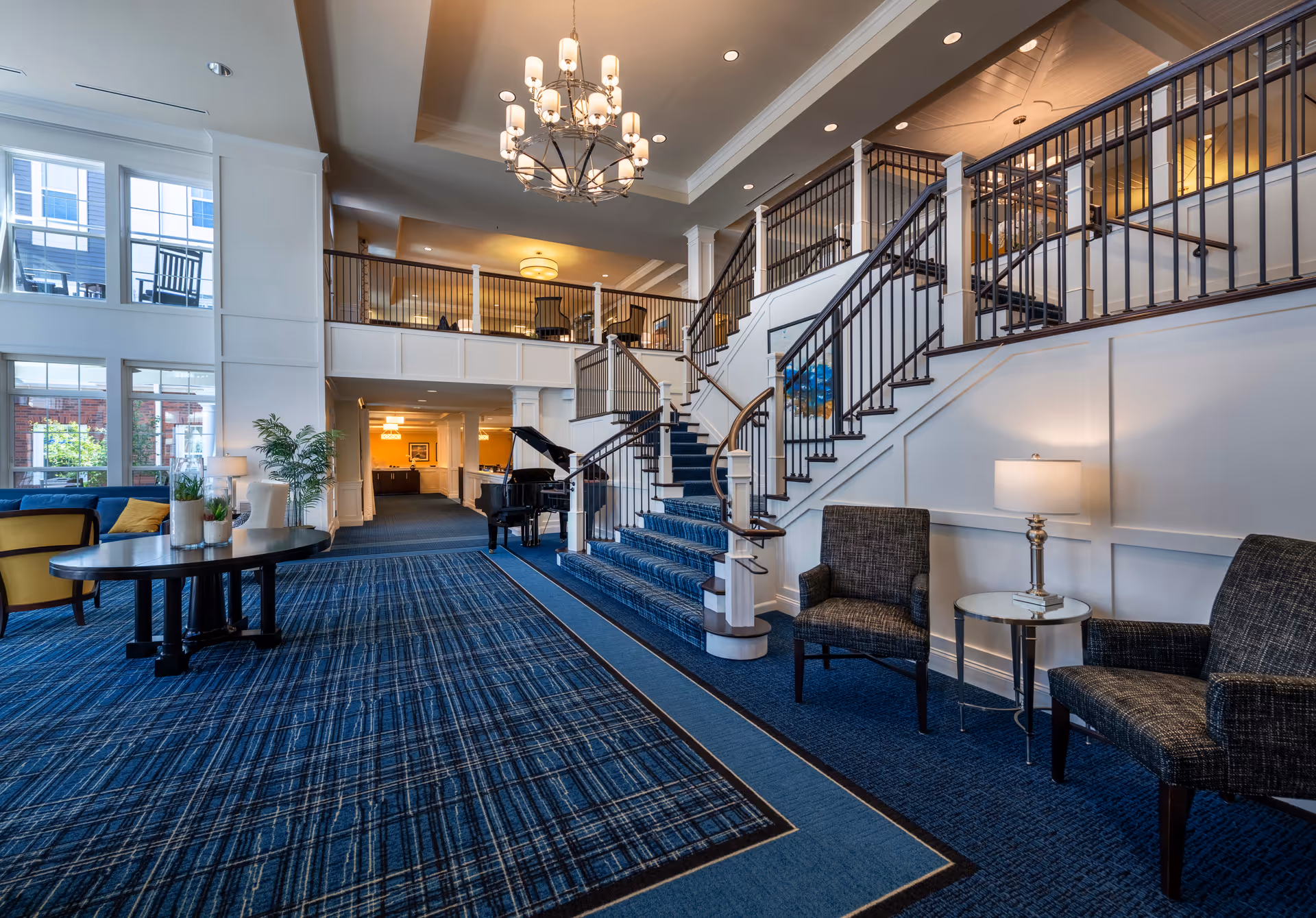 Spacious and elegant senior living facility common area with a blue patterned carpet, a grand staircase with dark railings, a black grand piano, comfortable seating including two armchairs and a small table with a lamp, large windows letting in natural light, and a chandelier hanging from the ceiling.
