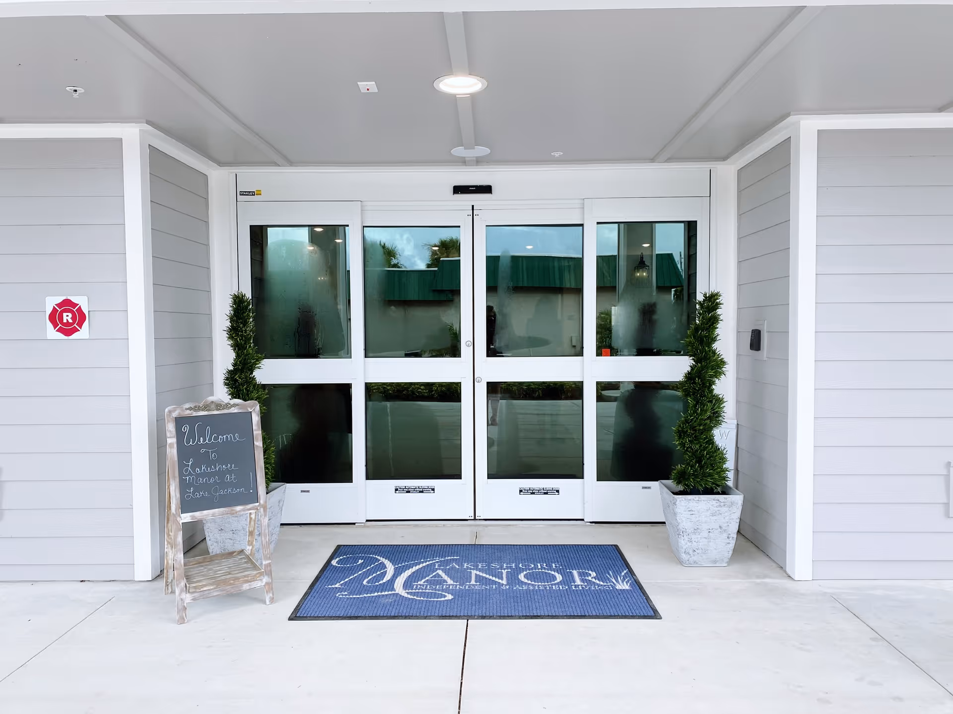 Front entrance of Lakeshore Manor with glass automatic doors, a welcome chalkboard, potted plants, and a blue entrance mat.