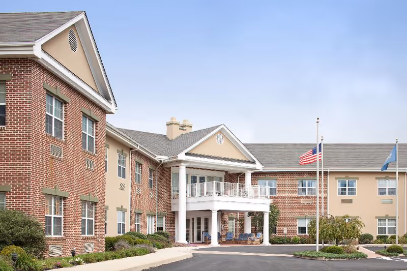 Exterior view of a two-story senior living facility building with a brick and beige facade, white columns supporting a covered entrance, and three flagpoles with flags in front of the building under a clear sky.