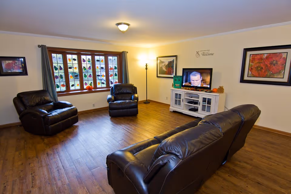 A cozy living room with wooden flooring, featuring three dark brown leather recliners arranged around a white TV stand with a flat-screen television. The room has a large window with colorful stained glass decorations and curtains, two framed floral paintings on the walls, a floor lamp in the corner, and small decorative pumpkins on the window sill and TV stand.