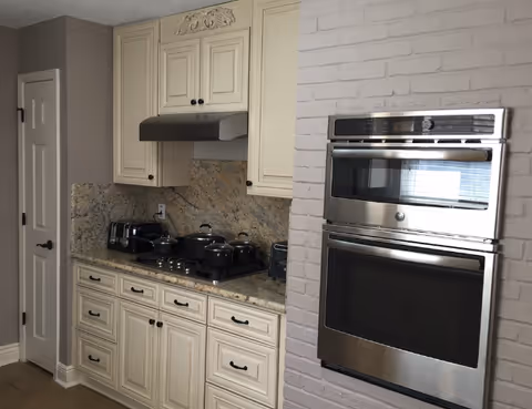 A kitchen area with cream-colored cabinets and granite countertops. There is a stovetop with several pots and pans, a toaster, and a toaster oven on the counter. To the right, there is a built-in stainless steel double oven set into a white brick wall. A closed white door is visible on the left side of the image.