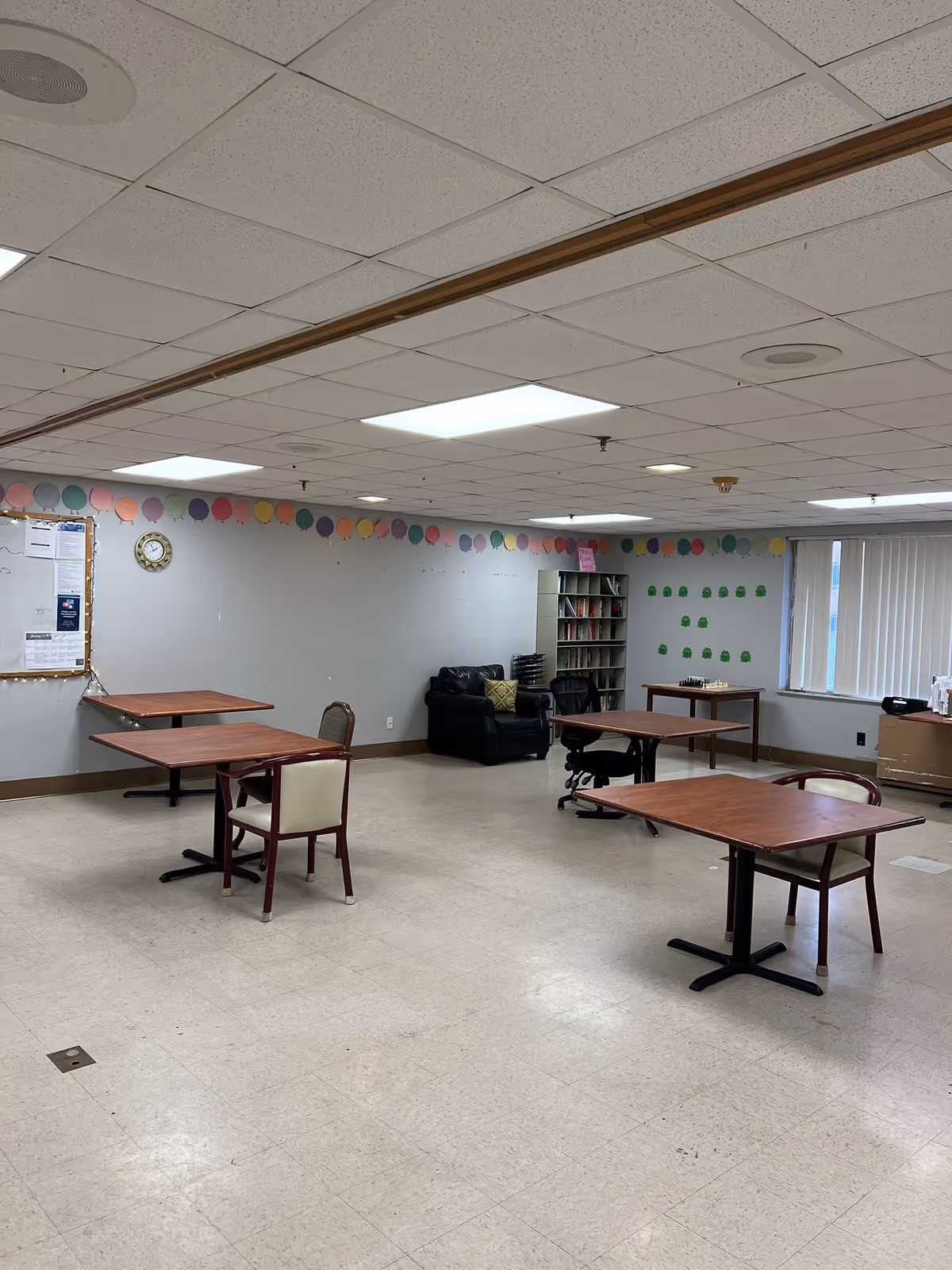 A spacious room with several wooden tables and chairs arranged sparsely. The walls are decorated with colorful circular cutouts near the ceiling and some green frog cutouts near a window with vertical blinds. There is a black leather armchair and a bookshelf filled with books in the corner. The ceiling has recessed lighting and a drop ceiling design.