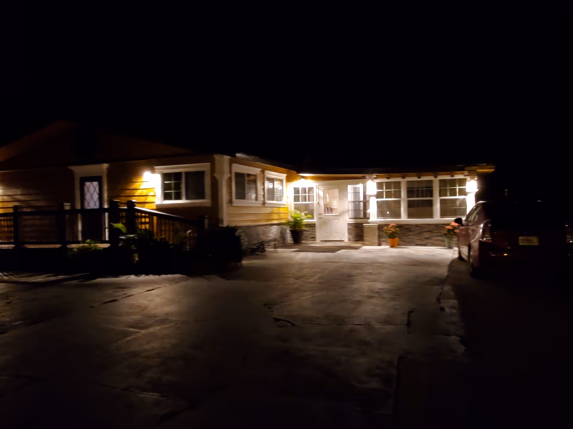 Night view of a single-story building with exterior lights illuminating the entrance and windows. There is a paved driveway in front with a parked car on the right side. The building has a porch area with a railing on the left and potted plants near the entrance.