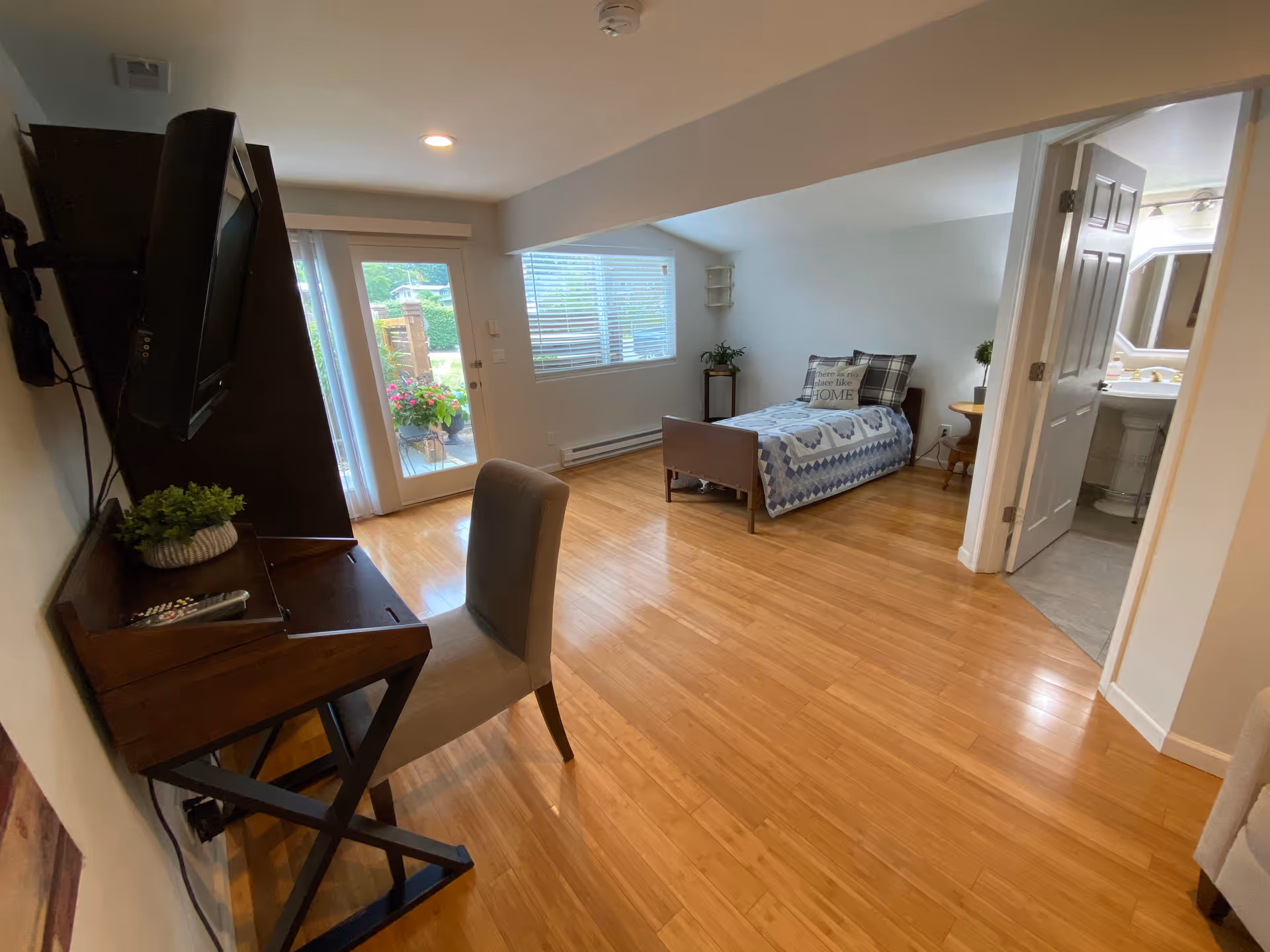 A senior living facility room with a single bed covered in a blue and white quilt, a pillow with the text 'There is no place like HOME', a small wooden side table with a plant, a desk with a chair and a mounted TV, wooden flooring, a door leading to a bathroom, and a glass door opening to an outdoor area with flowers.
