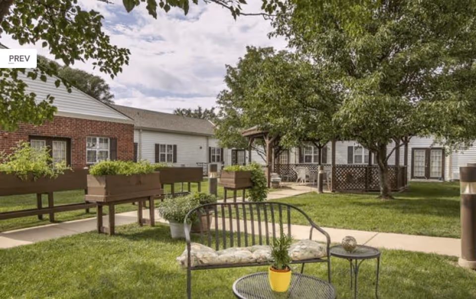 Outdoor garden area at Stratford Commons Memory Care Community with green grass, trees, raised planter boxes, a metal bench with cushions, and a small round table with a yellow potted plant. Residential-style buildings with white siding and brick accents surround the garden.