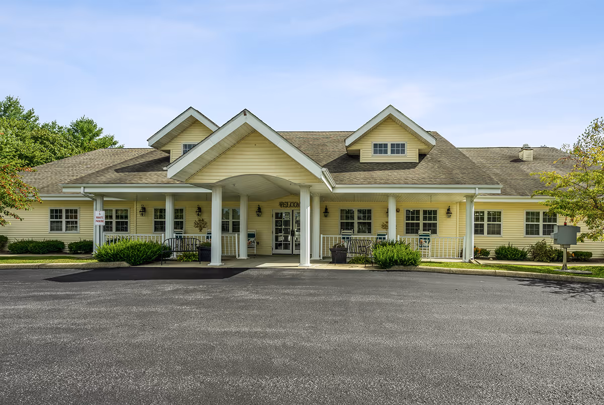Front exterior view of a single-story yellow building with a covered entrance supported by white columns, benches on the porch, and a paved driveway in front. The building is surrounded by greenery and trees under a clear sky.