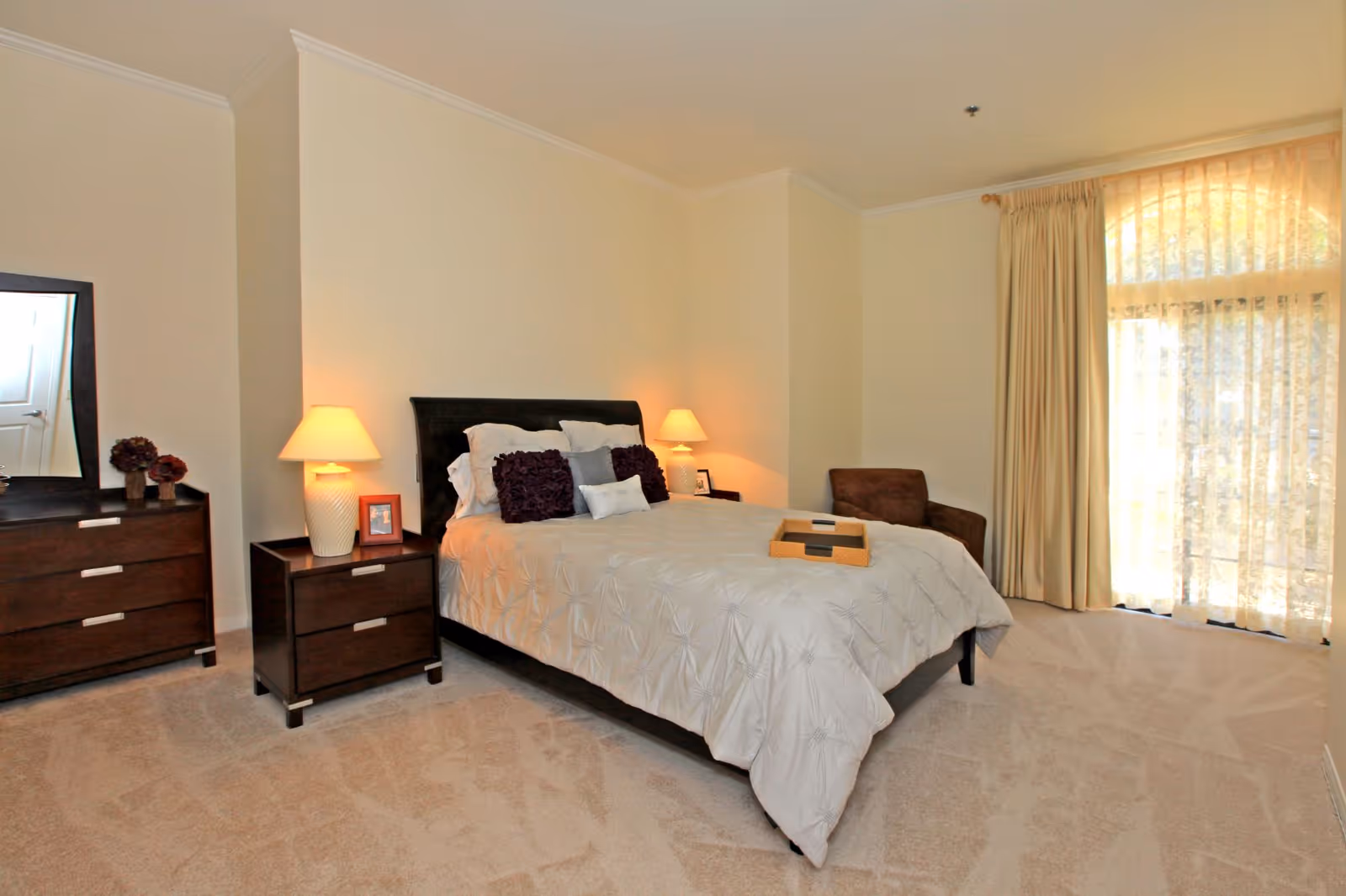 A well-lit bedroom with a large bed covered in a white textured comforter and several pillows. There are two dark wooden nightstands on either side of the bed, each with a lamp and framed photos. To the left, there is a matching dresser with a mirror and decorative items. A brown armchair is placed near a large window with sheer and heavy curtains, allowing natural light to fill the room. The carpet is light-colored and the walls are painted a soft cream.
