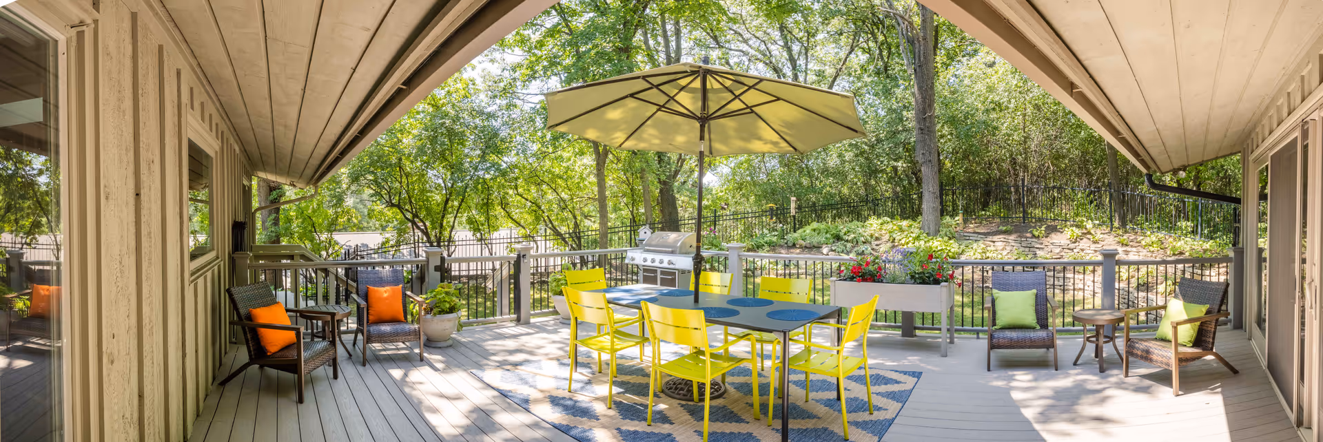 Covered outdoor deck with a yellow dining table and chairs under an umbrella, lounge seating, planters and a grill overlooking trees.
