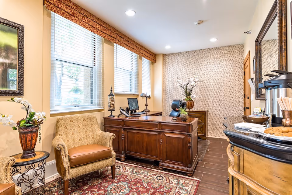 A warmly decorated office space with a wooden desk, a computer monitor, and a black office chair. There is a patterned armchair with a leather seat cushion next to a small round table holding a decorative plant. The room has three large windows with blinds and a valance, a patterned wallpaper accent wall, and a wooden floor with a red patterned rug. On the right side, there is a counter with a coffee dispenser, cups, and cookies. A large mirror hangs on the wall above the counter.
