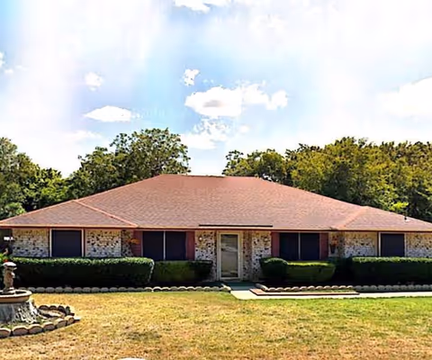 Single-story brick building with a red roof, surrounded by trimmed bushes and trees in the background under a partly cloudy sky.