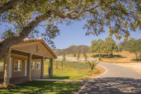 A sunny outdoor scene at Villa Monticello featuring a building with a covered porch supported by columns on the left, a winding paved road leading uphill, green grass, trees, and a white fence with mountains in the background under a clear blue sky.