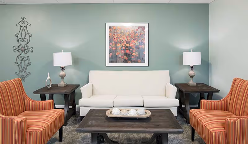 A cozy living room with a white three-seat sofa centered against a light green wall. On either side of the sofa are dark wooden side tables, each with a lamp and decorative items. Two striped orange armchairs face the sofa, and a dark wooden coffee table with decorative spheres sits in front. A colorful floral painting hangs above the sofa.