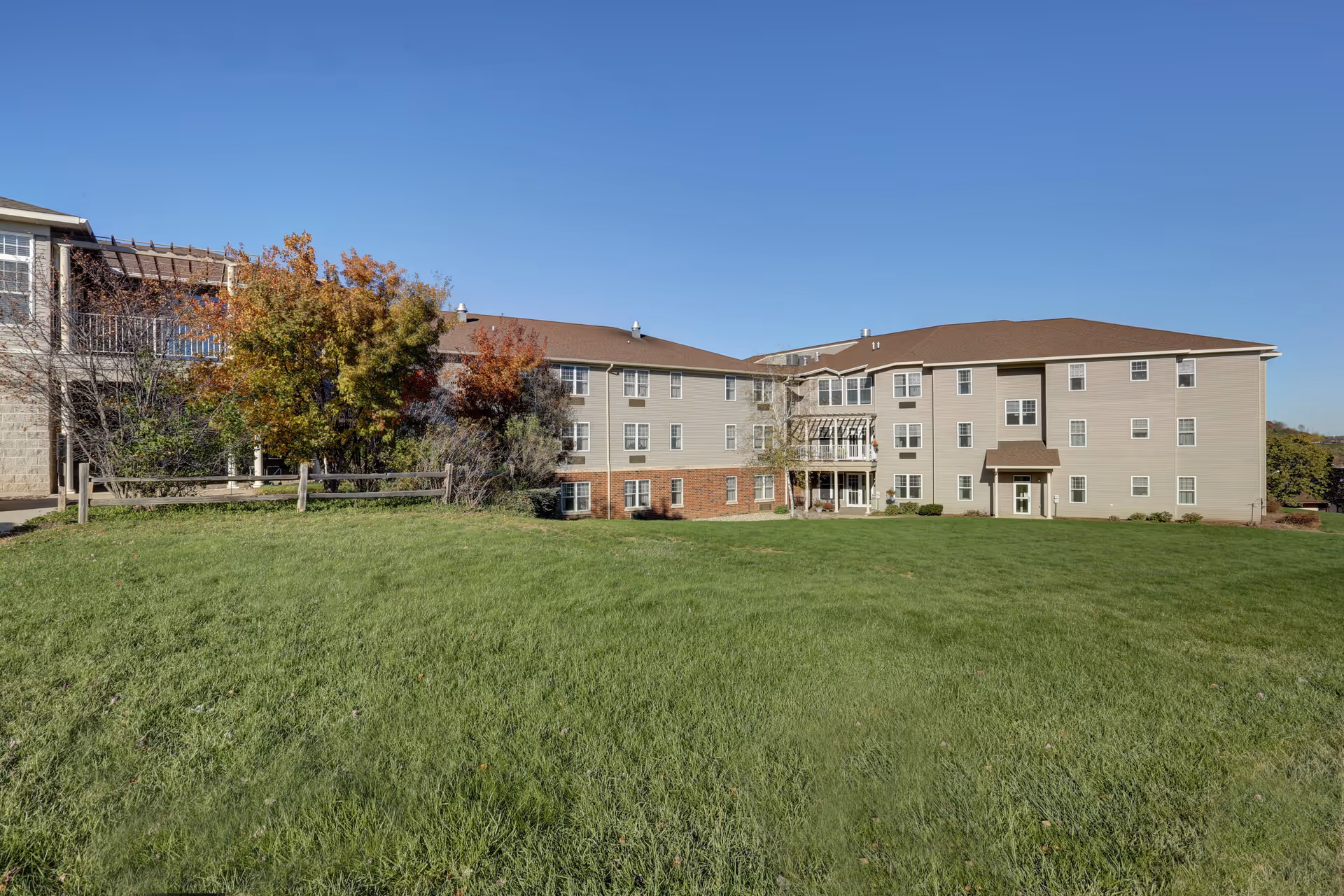 Three-story senior living building with a large green lawn and trees under a clear blue sky.