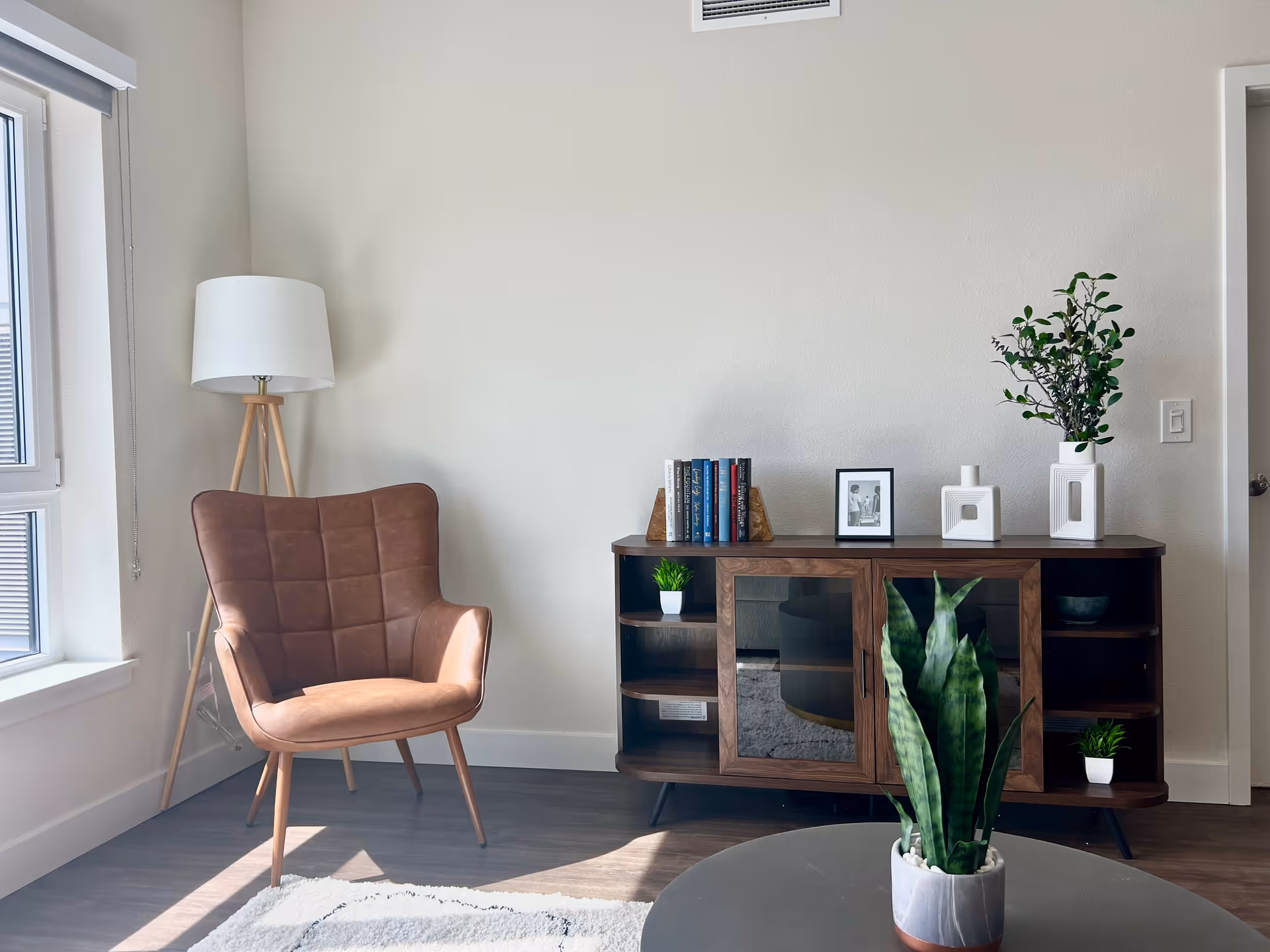 A cozy corner of a living room featuring a brown cushioned armchair with wooden legs next to a tall tripod floor lamp with a white lampshade. Adjacent to the chair is a wooden sideboard with glass doors, decorated with books, a small framed photo, white ceramic vases, and green plants. In the foreground, there is a round table with a potted snake plant. The room has light-colored walls, a window with blinds, and wooden flooring.