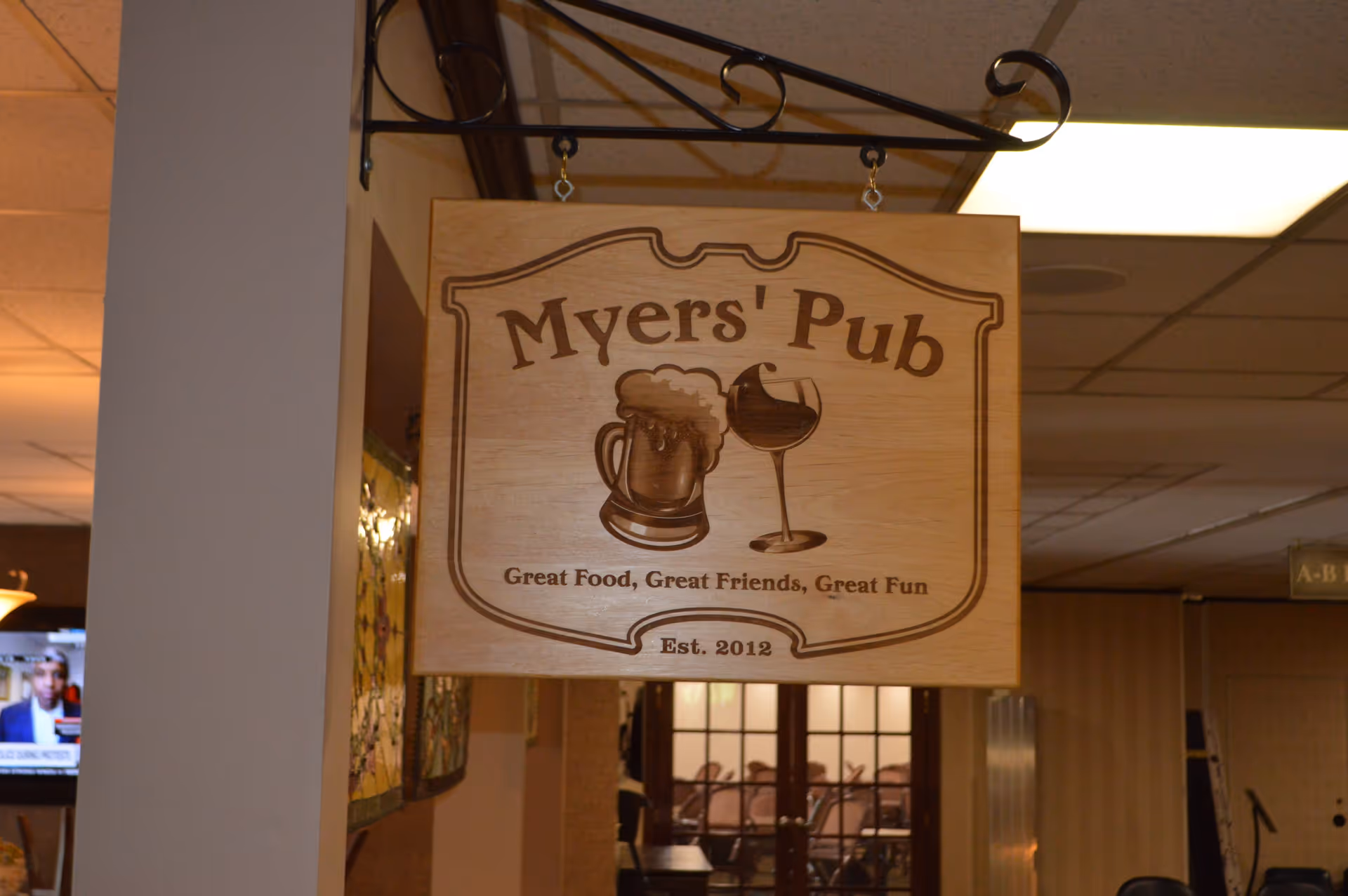 A wooden hanging sign inside a building that reads 'Myers' Pub' with images of a beer mug and a wine glass. Below the name, the sign says 'Great Food, Great Friends, Great Fun' and 'Est. 2012'. The background shows part of an interior hallway with a television and some chairs visible.