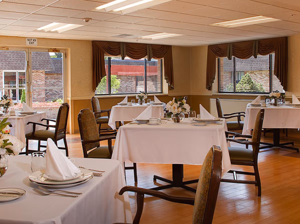 Bright dining room with tables dressed in white tablecloths, place settings, floral centerpieces, and chairs near windows.