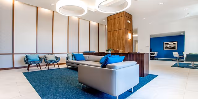 Modern lounge with gray sofas and blue accent pillows, chairs, circular ceiling lights, and a blue area rug in a bright common room.