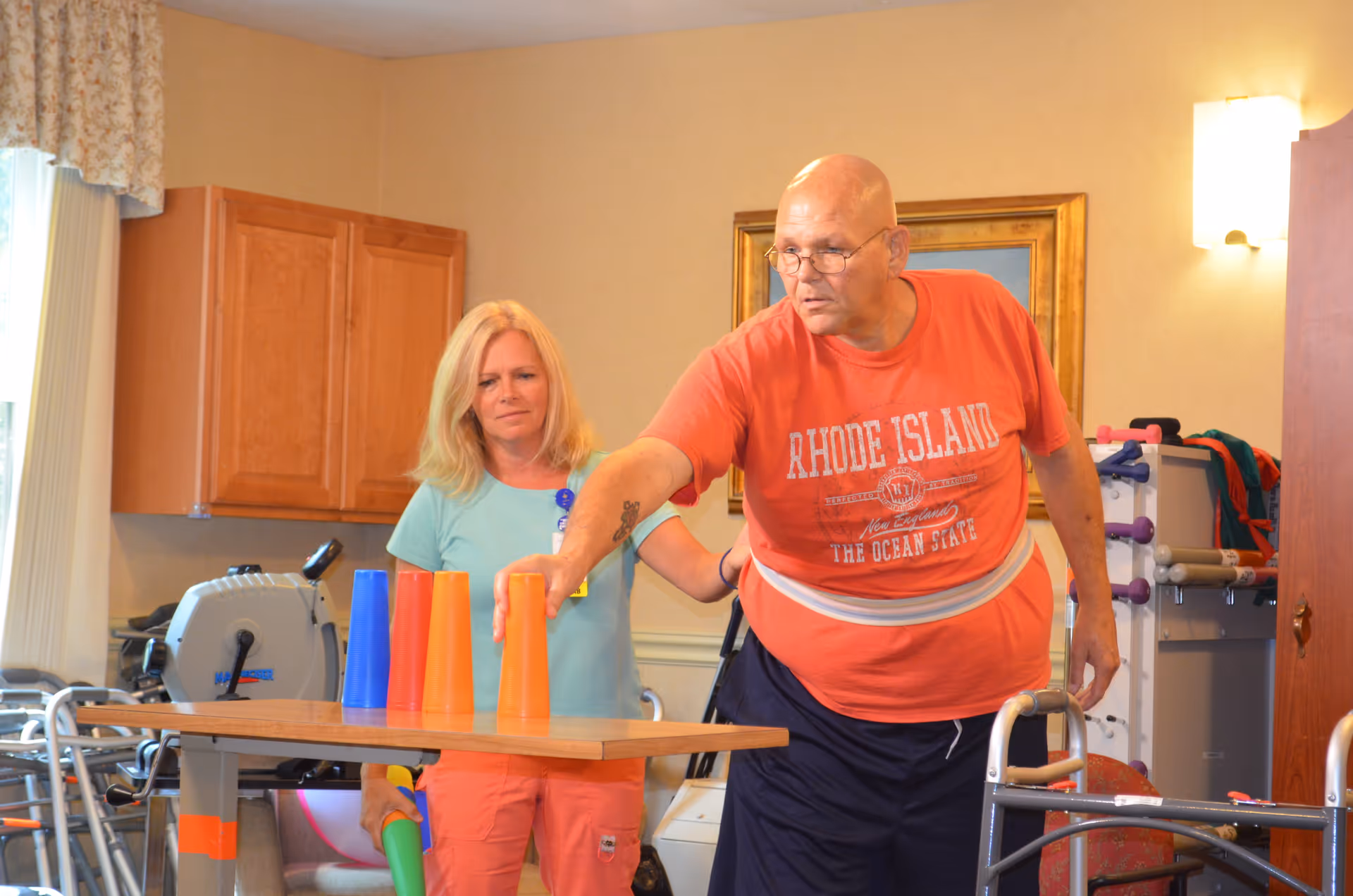 A man wearing a red Rhode Island t-shirt is stacking colorful plastic cups on a table while a woman in a light blue shirt watches and assists him in a room with exercise equipment and wooden cabinets.