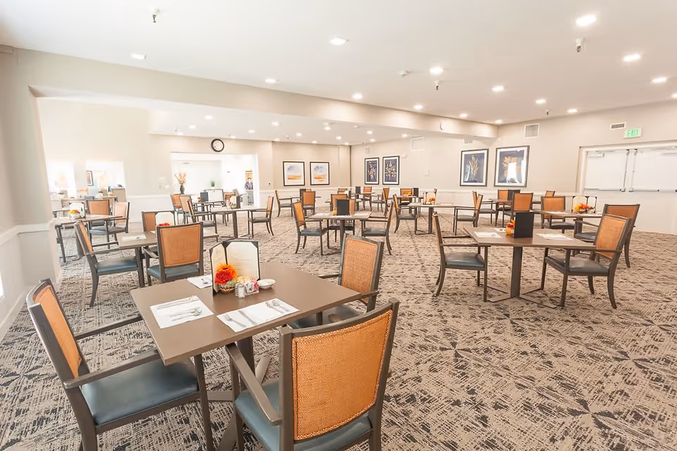 Spacious dining room with multiple square tables and chairs arranged neatly. Each table is set with placemats, utensils, and small floral centerpieces. The room has a patterned carpet, light-colored walls, framed artwork, and bright ceiling lights.