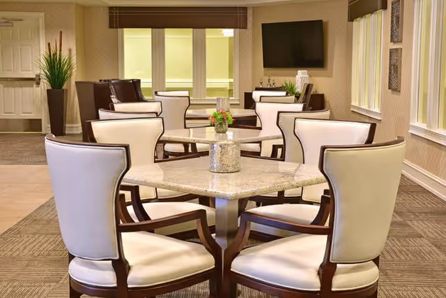 A well-lit dining area with multiple square tables surrounded by cream-colored cushioned chairs with dark wooden frames. The room features beige walls, large windows with blinds, a flat-screen TV mounted on the wall, and decorative plants and vases on tables and furniture.