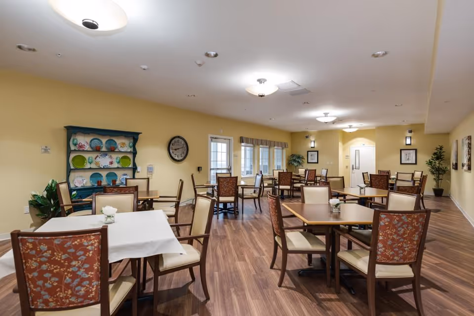 A spacious dining room with multiple tables and chairs arranged neatly. The walls are painted yellow and decorated with framed pictures and a clock. A blue hutch with colorful plates is visible on the left side. The room has wooden flooring and ceiling lights providing bright illumination.