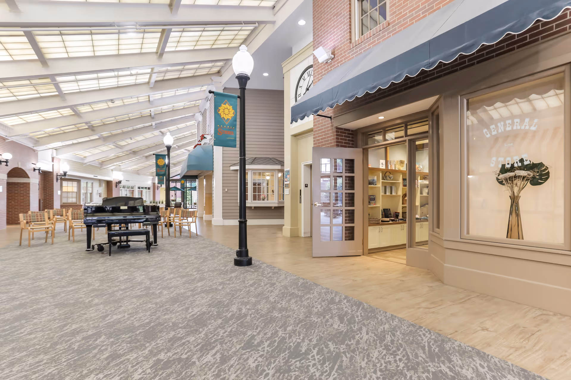 Indoor common area with a high ceiling and large skylights, featuring a black grand piano surrounded by wooden chairs. There are streetlamp-style lights and banners along the walkway. On the right side, there is a storefront with a sign that reads 'General Store' and an open door showing shelves with various items inside.