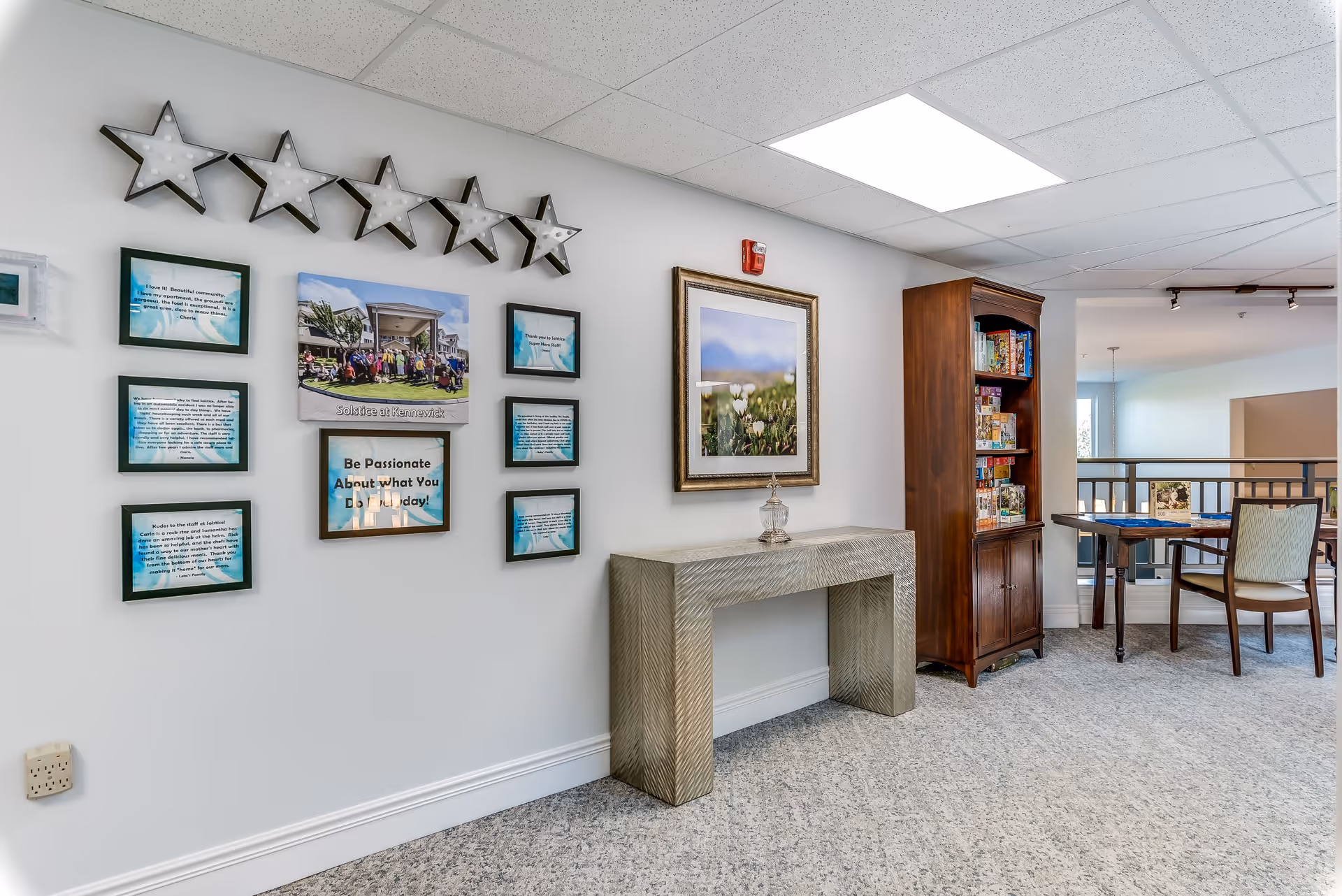 An interior common area in a senior living facility featuring a wall decorated with framed photos and quotes, illuminated star-shaped wall lights, a textured console table with a decorative glass item, a wooden cabinet filled with board games, and a table with chairs near a railing overlooking a lower level.