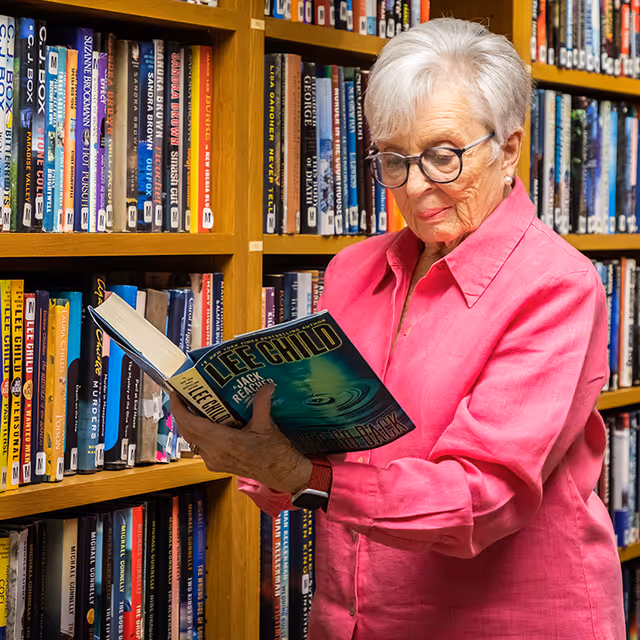 An elderly woman with short white hair and glasses is standing in front of a bookshelf filled with books. She is wearing a bright pink shirt and is reading a book titled 'Jack Reacher' by Lee Child.
