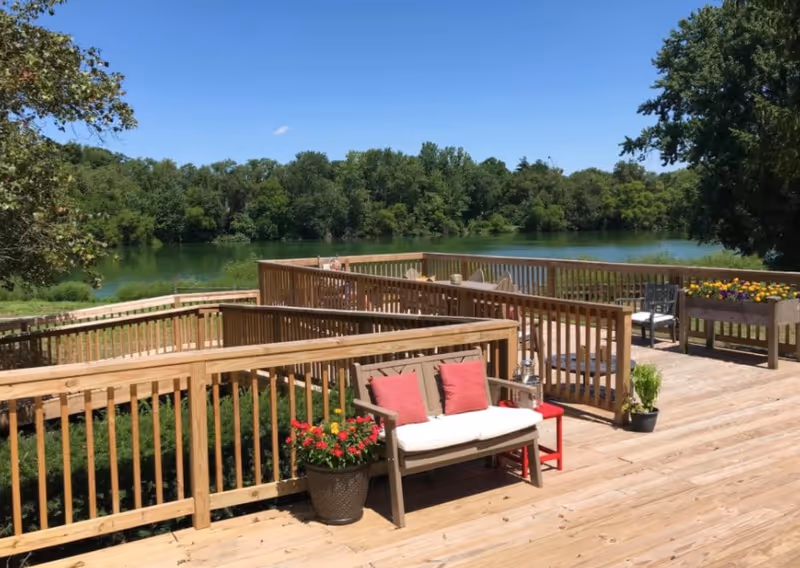 A wooden deck with railings overlooking a calm lake surrounded by trees under a clear blue sky. The deck has a bench with red cushions, potted plants, and a small red side table. There are also flower boxes with colorful flowers and additional seating further along the deck.