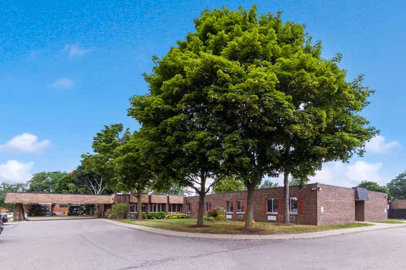 Single-story brick senior living facility with a covered entrance, large trees and a driveway under a blue sky.