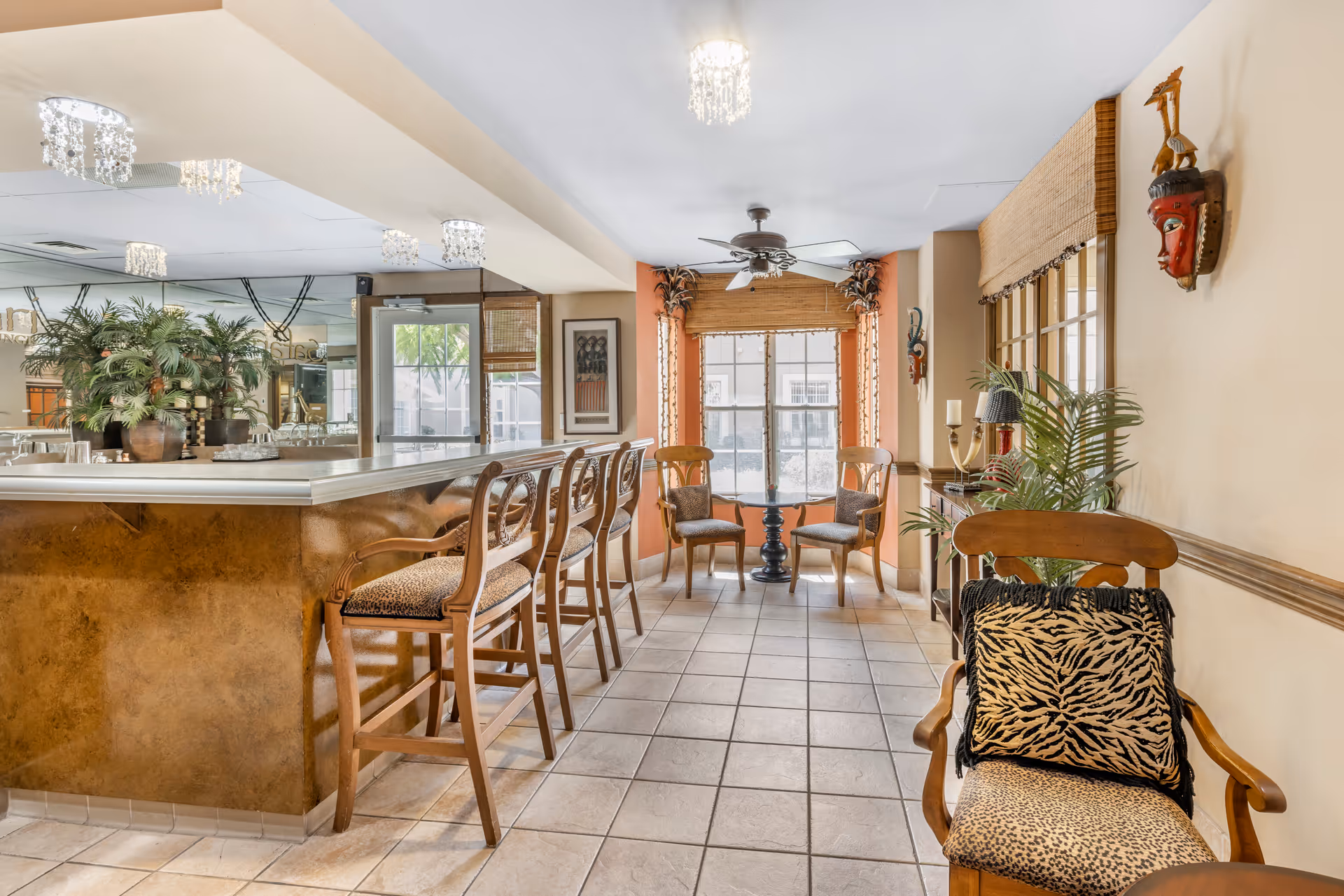 Interior view of a lounge area in Brookdale Mirage Inn featuring a bar counter with four wooden bar stools upholstered in animal print fabric. There are two wooden chairs with cushions near a window with bamboo blinds, a ceiling fan, decorative plants, and wall-mounted masks. The floor is tiled and the lighting includes small chandeliers.