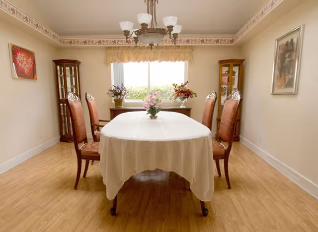 A dining room with a rectangular table covered with a white tablecloth and surrounded by six wooden chairs with red upholstery. The room has light-colored walls with floral border trim near the ceiling, a wooden floor, and a window with a yellow valance letting in natural light. There are two glass display cabinets on either side of the window and floral arrangements on the table and window sill. A chandelier with multiple lights hangs above the table.