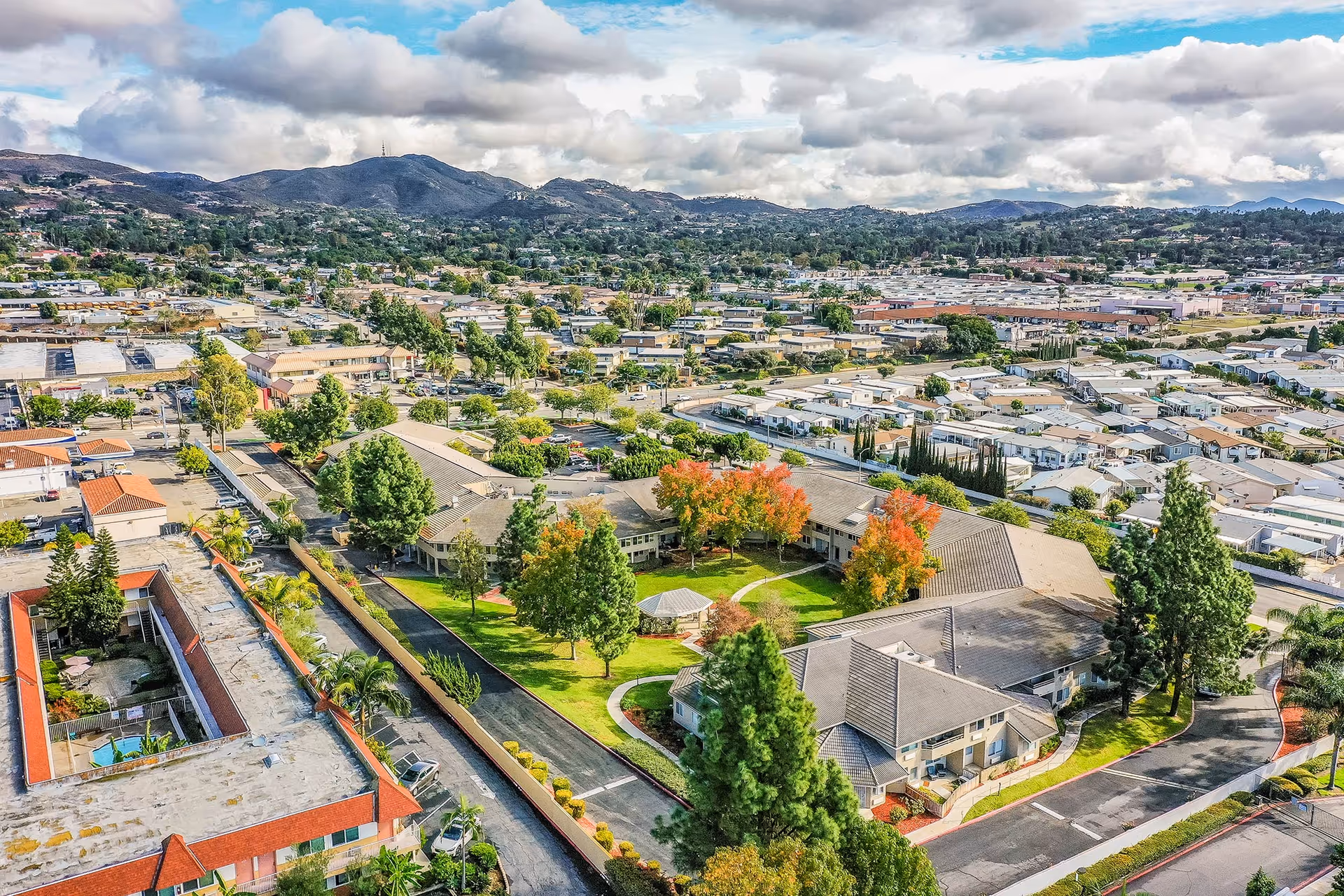 Aerial view of a senior living facility surrounded by greenery and trees with some showing autumn colors. The facility buildings are arranged around a central courtyard with a gazebo. The surrounding area includes residential and commercial buildings with hills and mountains in the background under a partly cloudy sky.