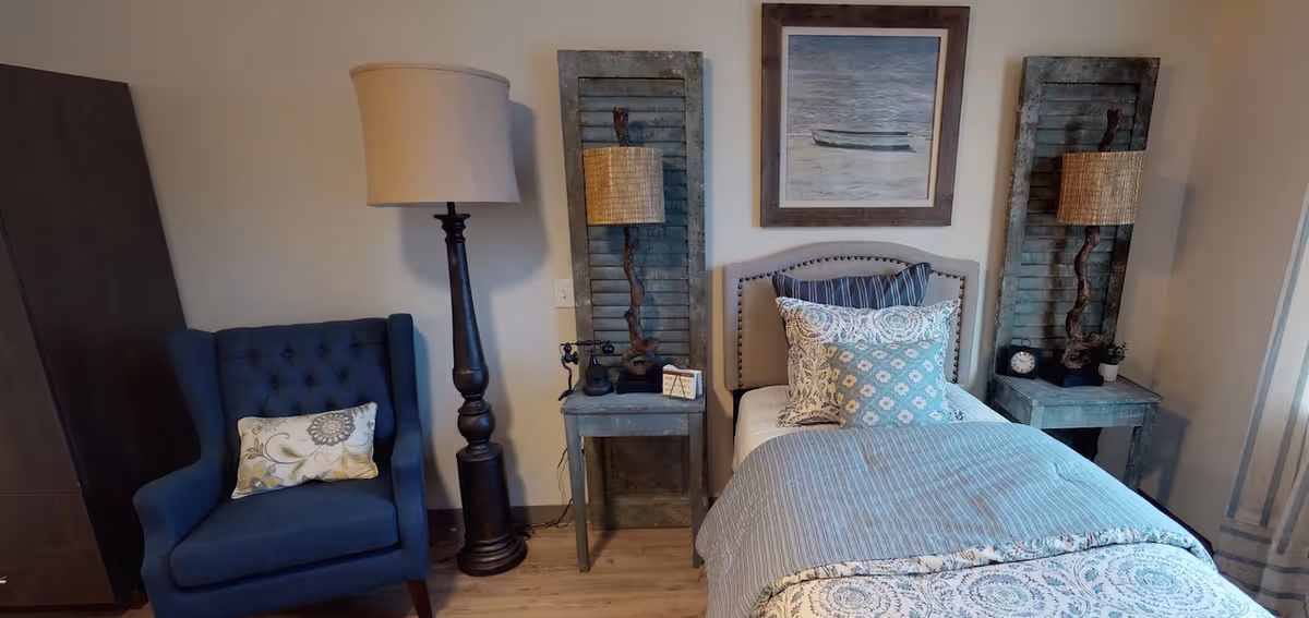 A cozy bedroom featuring a single bed with patterned bedding and multiple pillows, flanked by two rustic nightstands each holding a unique lamp. To the left of the bed is a tall floor lamp and a dark blue upholstered armchair with a decorative pillow. The walls are light-colored, adorned with a framed picture above the bed and two wooden shutter panels behind the nightstands.
