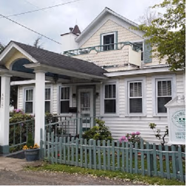 Exterior view of a two-story residential building with white siding and a small front porch. The porch has a green railing and a ramp leading to the entrance. There is a green picket fence in front with some flowering plants and shrubs. A sign is visible near the fence, partially readable, indicating it is a facility named Good Living.